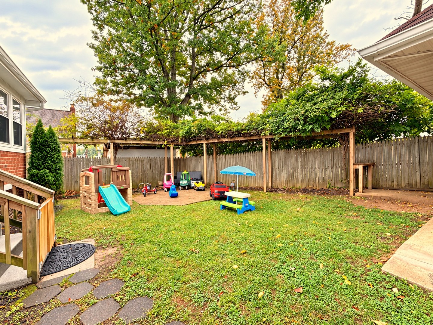 Fenced backyard play yard with playhouse, slide, ride-on toys, and picnic table
